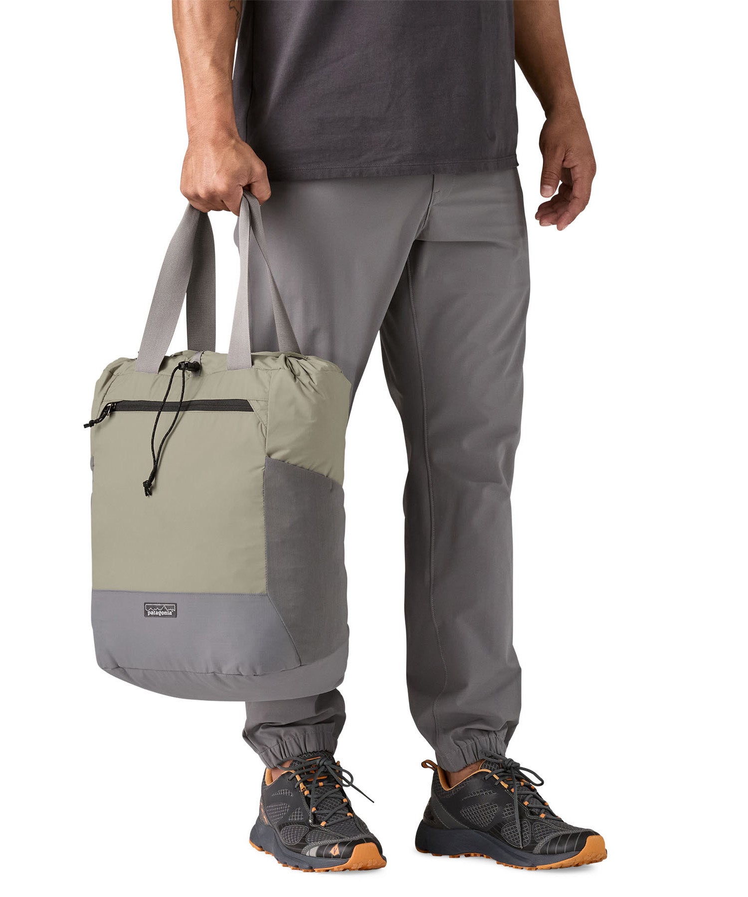 A close up of the Patagonia Terravia tote bag in river rock green carried by an adult by the handles showing the front zip details and 2 side pockets.