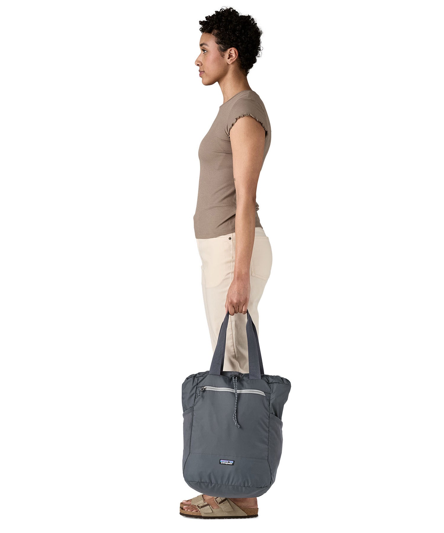 A side view of the Patagonia Terravia tote bag in smolder blue carried by an adult by the handles showing the front zip details and 2 side pockets.