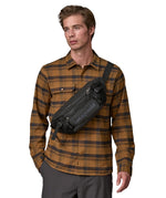 A man wearing the patagonia waist pack in black over the shoulder. A 100% recycled material pack with 2 main compartments and adjustable shoulder strap.