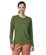 A woman wearing a Patagonia Women's long sleeved Cap Cool Daily Graphic Shirt in a Green X-Dye colour. The woman is stood in front of a plain background. 