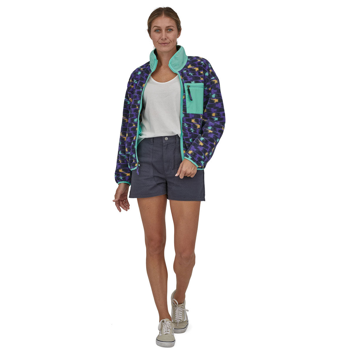 Woman wearing the Patagonia women's synchilla fleece in perennial purple with the zip fully open, with purple shorts, a white t-shirt and trainers. 