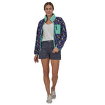 Woman wearing the Patagonia women's synchilla fleece in perennial purple with the zip fully open, with purple shorts, a white t-shirt and trainers. 
