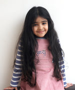 A child wearing the Pigeon Organics pink corduroy pinafore dress with a pink cat appliqué on the front pocket