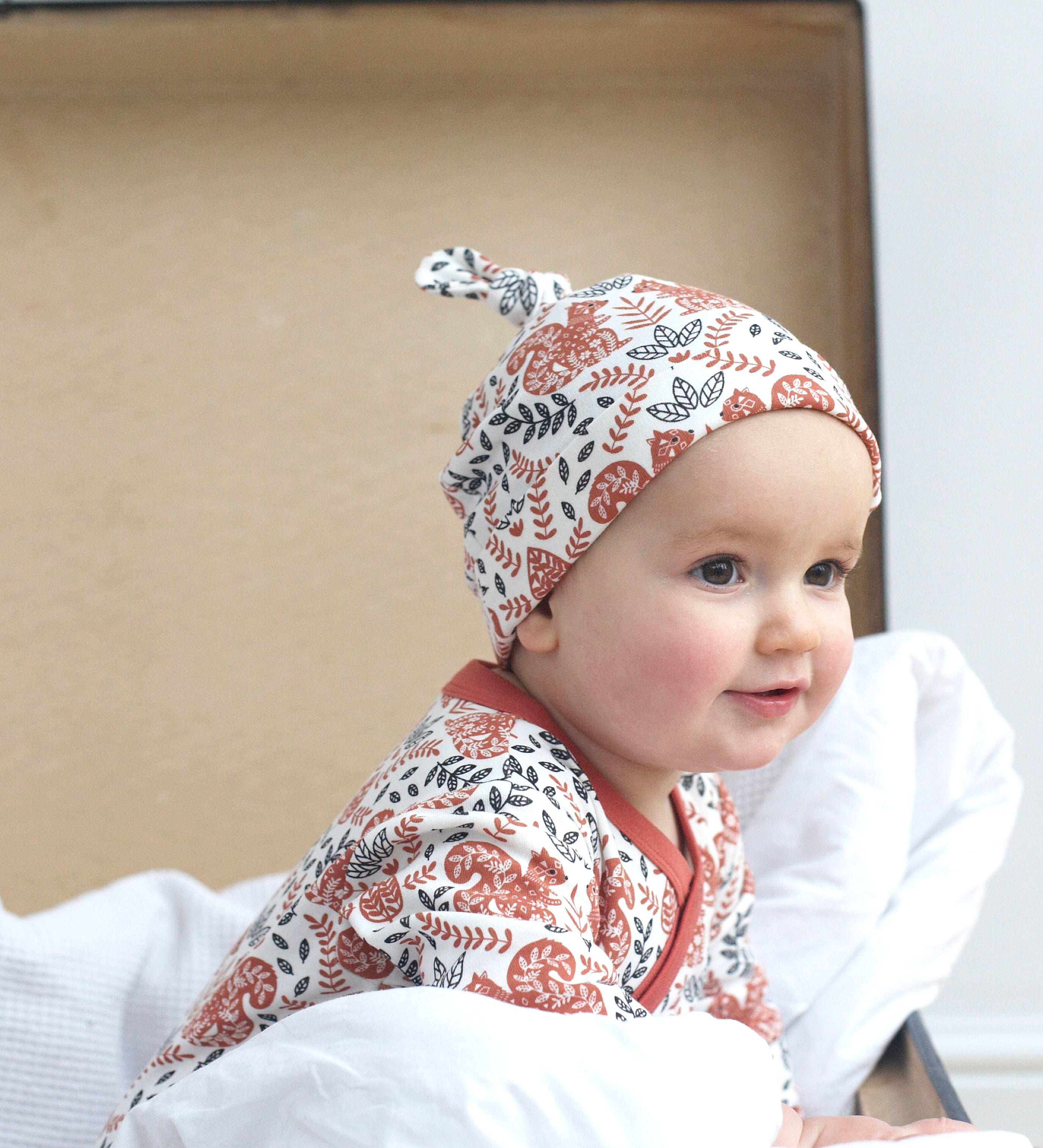 A close up of a baby sitting inside a trunk wearing the Pigeon Organics kimono romper and the knotted hat. Showing the squirrel and floral print on organic cotton fabric