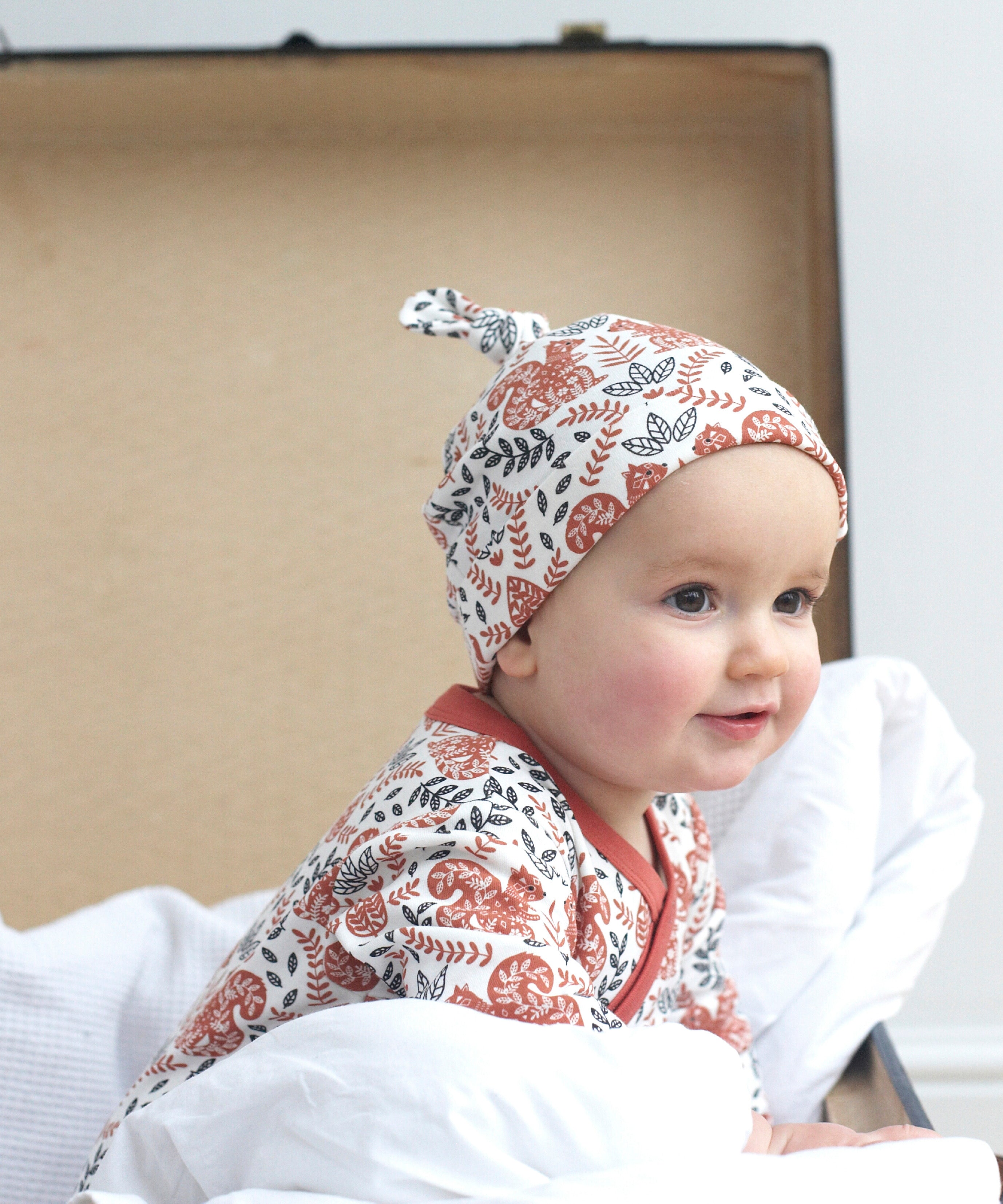 A close up of a baby sitting inside a trunk wearing the Pigeon Organics kimono romper and the knotted hat. Showing the squirrel and floral print on organic cotton fabric