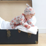 A baby playing inside a trunk on a wooden surface wearing the wearing the Pigeon Organics kimono romper and the knotted hat. Showing the squirrel and floral print on organic cotton fabric