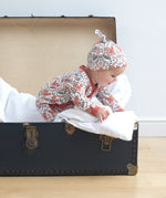 A baby playing inside a trunk on a wooden surface wearing the wearing the Pigeon Organics kimono romper and the knotted hat. Showing the squirrel and floral print on organic cotton fabric