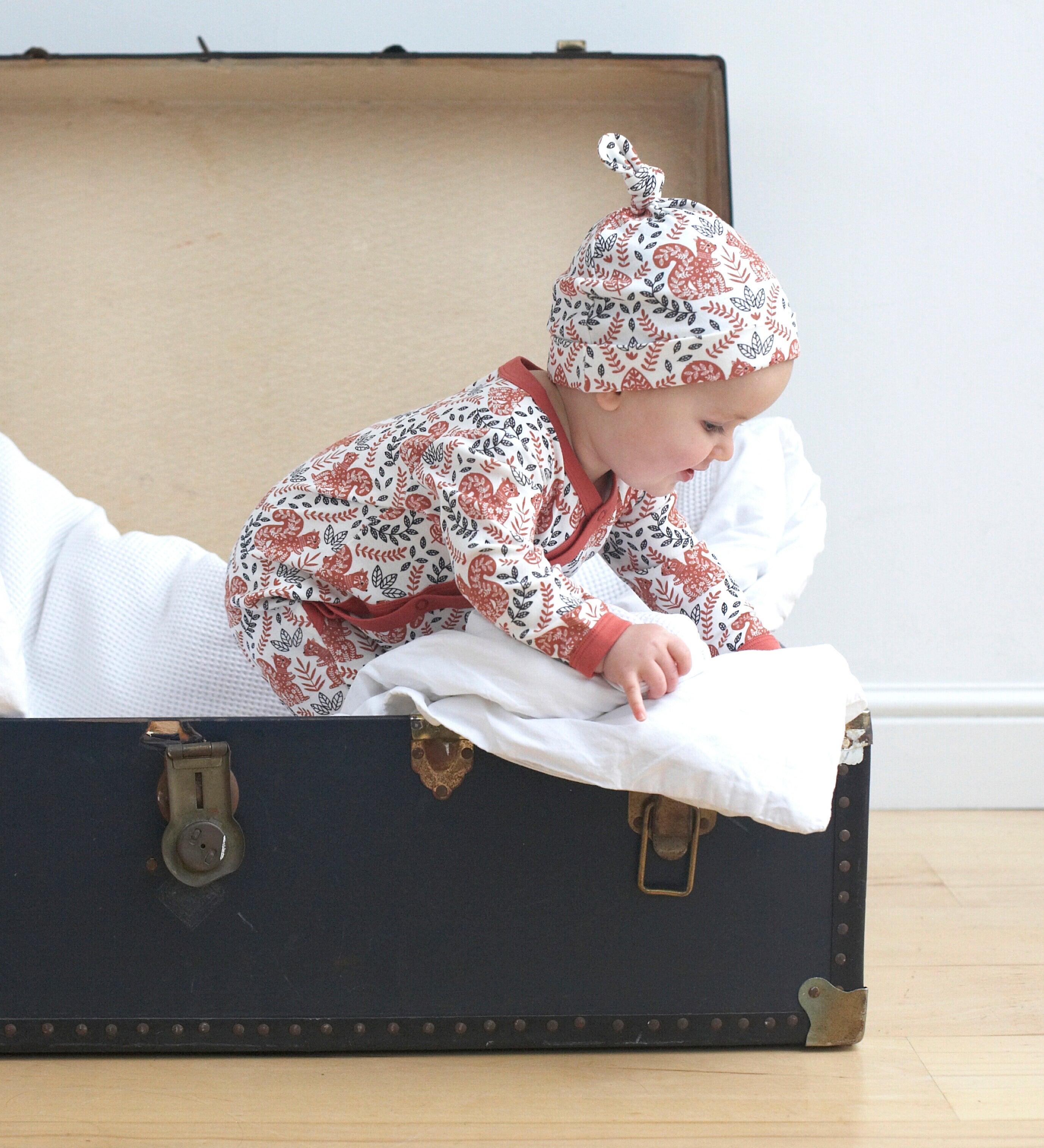 A baby playing inside a trunk on a wooden surface wearing the wearing the Pigeon Organics kimono romper and the knotted hat. Showing the squirrel and floral print on organic cotton fabric