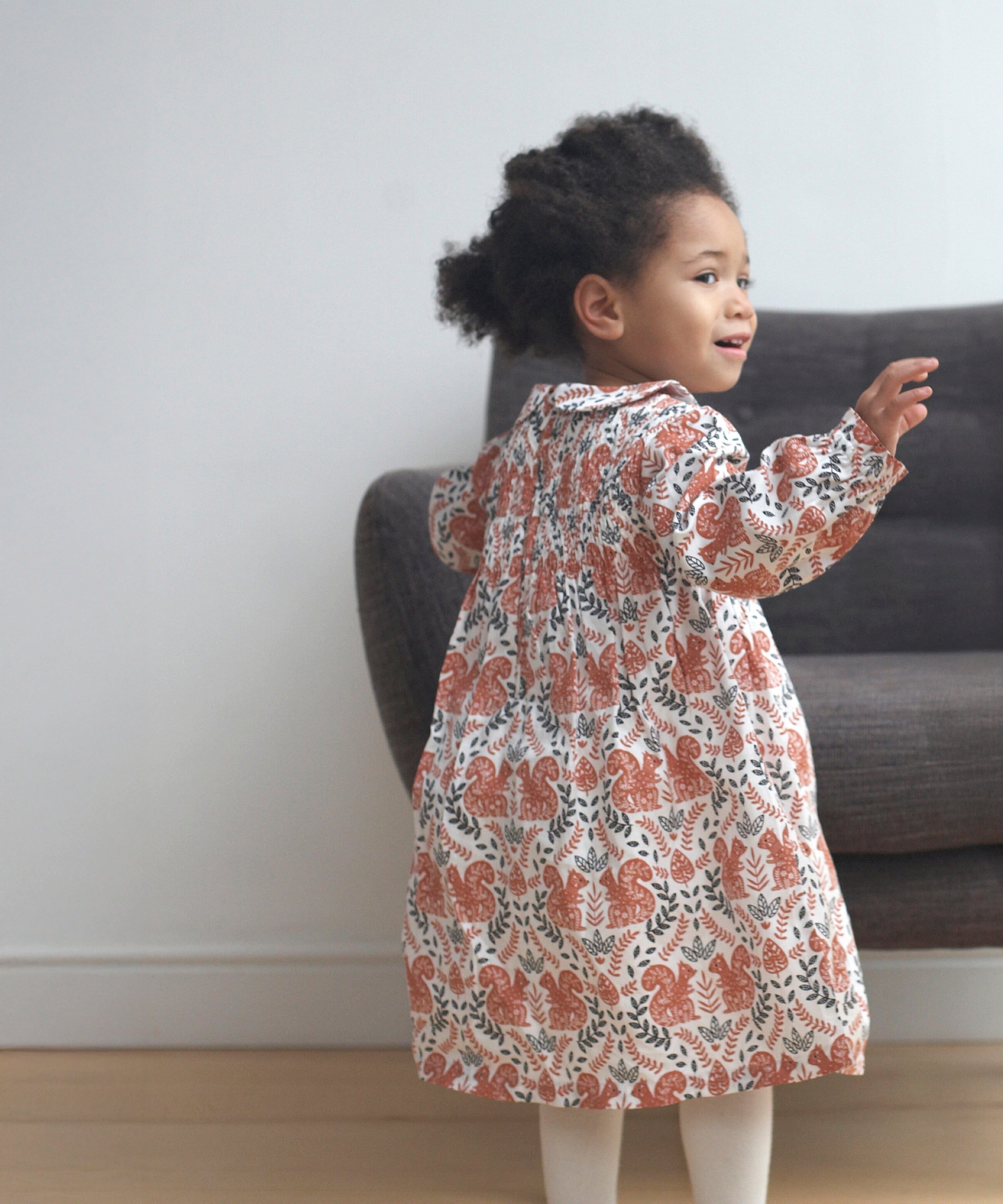 A child wearing the Pigeon Organics squirrel smock dress standing on a wooden surface showing the curved Peter Pan style collar and elasticated waist band detail
