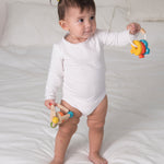 A baby standing on as bed holding onto the PlanToys Baby Key Rattle in one hand and the PlanToys Baby triangle clutching toy in the other. 


