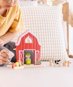 Child playing with the PlanToys barn house small world play set. Red wooden barn, mini veg patch, fences, cow, sheep, hen and farmer figure. 