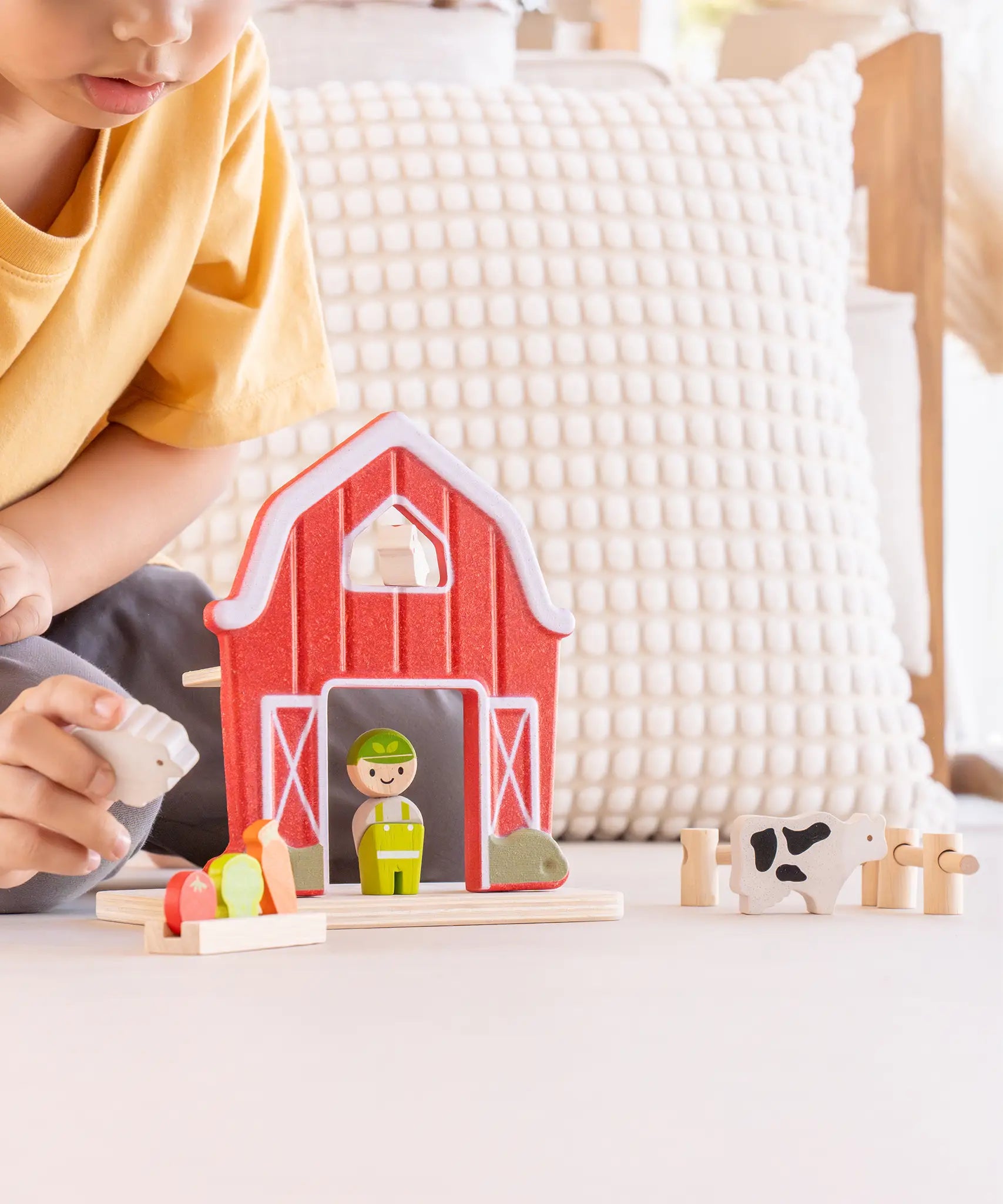 Child playing with the PlanToys barn house small world play set. Red wooden barn, mini veg patch, fences, cow, sheep, hen and farmer figure. 
