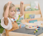 Child holding up a neckalace created with a white lace filled with beads from the PlanToys threading beads wooden lacing toy set. The wooden storage box is in front of them on a PlanToys children's desk. 