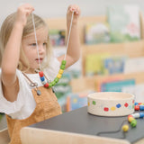 Child holding up a neckalace created with a white lace filled with beads from the PlanToys threading beads wooden lacing toy set. The wooden storage box is in front of them on a PlanToys children's desk. 