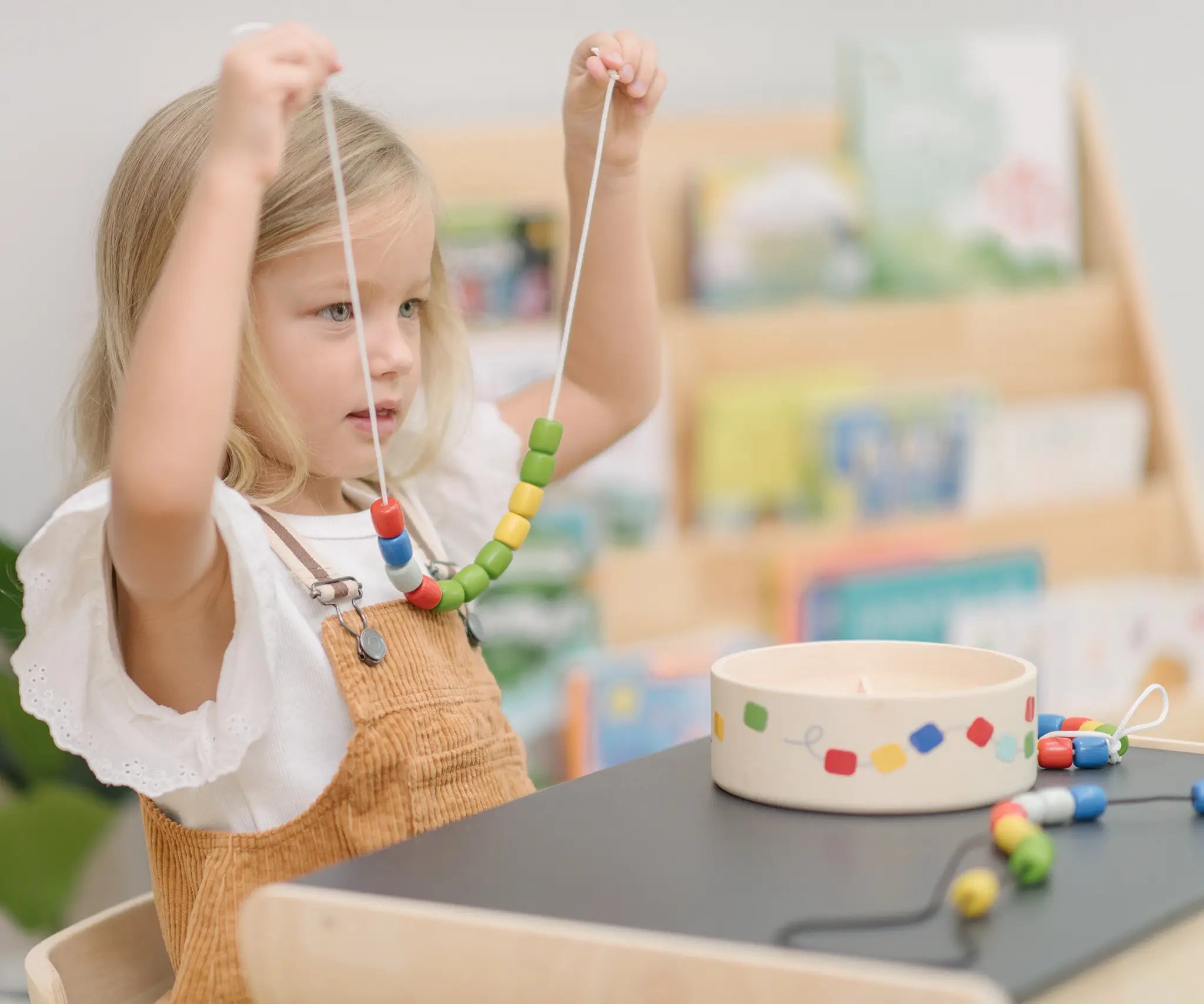 Child holding up a neckalace created with a white lace filled with beads from the PlanToys threading beads wooden lacing toy set. The wooden storage box is in front of them on a PlanToys children's desk. 