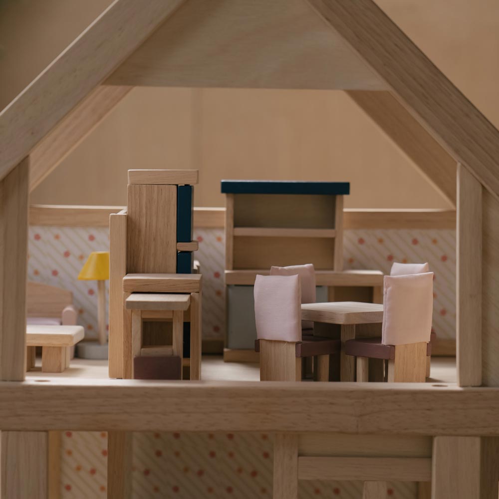 A side on view of the pieces from the PlanToys Dining Room Dolls House Furniture set set up in a wooden dolls house. 
