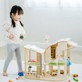 A child standing besides the PlanToys Green Dolls' House and Furniture Set. This eco inspired house has a lot of earth friendly features like a pretend recycling point, wind turbine, solar panel and grass and plants. 