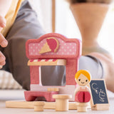 Close up of a child's hand reaching the wooden doll seated on one of the toy stools from the PlanToys ice cream shop wooden toy playset.