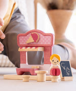 Close up of a child's hand reaching the wooden doll seated on one of the toy stools from the PlanToys ice cream shop wooden toy playset.