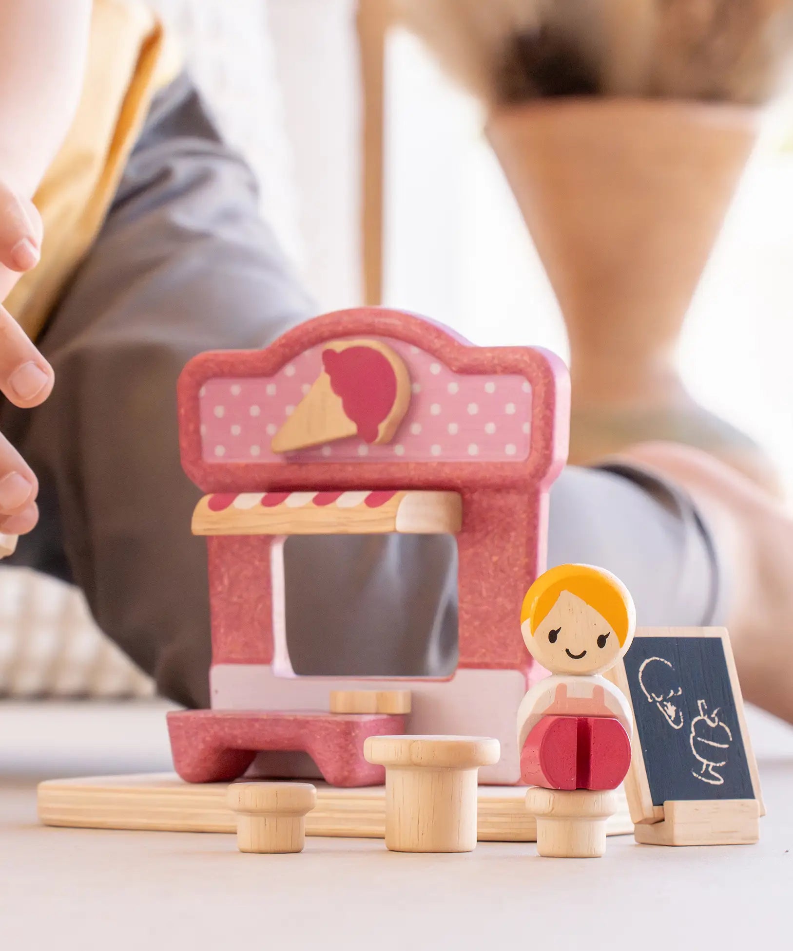 Close up of a child's hand reaching the wooden doll seated on one of the toy stools from the PlanToys ice cream shop wooden toy playset.