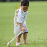 A child playing with a the PlanToys Mini Golf Set outside on grass. He is holding one of the clubs taking a swing at one of the balls. He has the canvas storage bag on his back with the other club poking out.