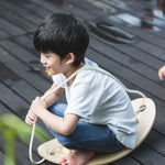 A child sitting on the PlanToys Balance Board holding the rope that is attached to the board. 