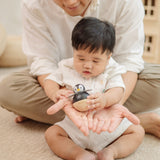 A baby sitting in front of an adult. The adult is handing a PlanToys Penguin Wobbler wooden baby toy to them. A circular shaped penguin toy that wobbles when placed on a flat surface. PlanToys range of toys are made from sustainably sourced rubberwood, we're proud stockists of PlanToys here at Babipur. 