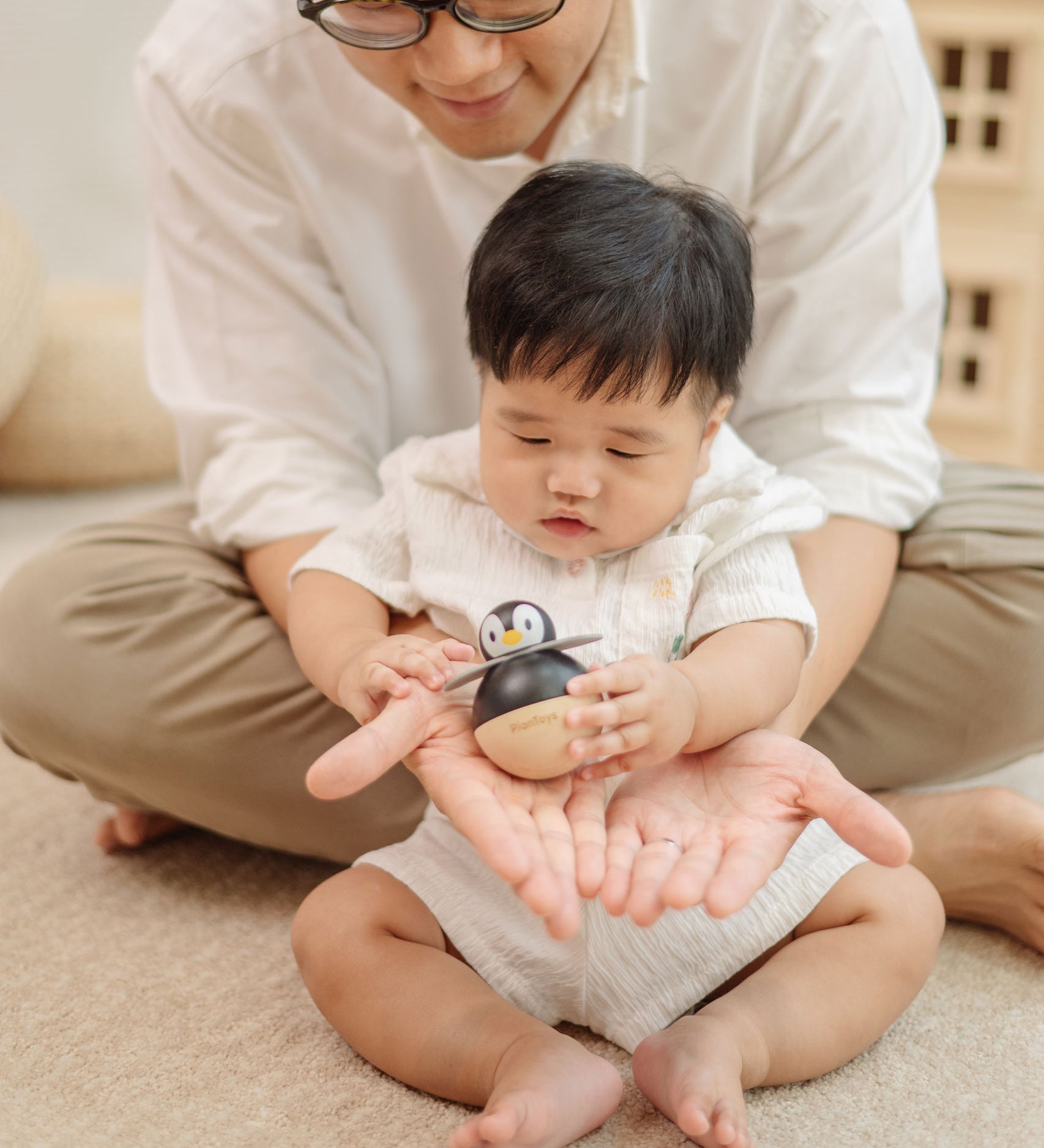 A baby sitting in front of an adult. The adult is handing a PlanToys Penguin Wobbler wooden baby toy to them. A circular shaped penguin toy that wobbles when placed on a flat surface. PlanToys range of toys are made from sustainably sourced rubberwood, we're proud stockists of PlanToys here at Babipur. 