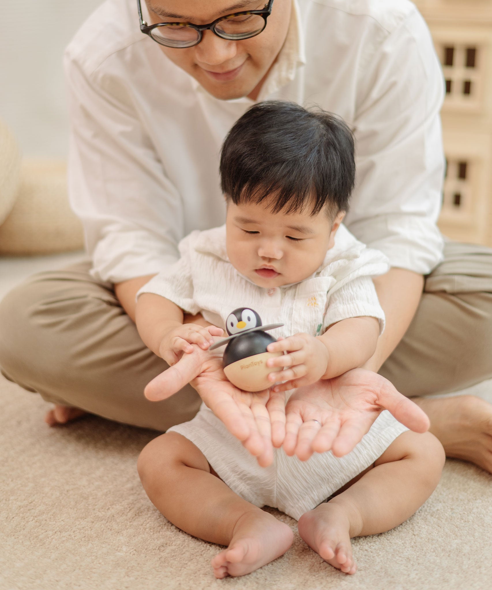 A baby sitting in front of an adult. The adult is handing a PlanToys Penguin Wobbler wooden baby toy to them. A circular shaped penguin toy that wobbles when placed on a flat surface. PlanToys range of toys are made from sustainably sourced rubberwood, we're proud stockists of PlanToys here at Babipur. 