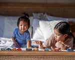 Two children playing with PlanToys vehicles whilst laying on their fronts on a bed. 