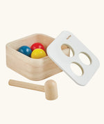 PlanToys punch and drop bin wooden pounding toy. Wooden box with three slots on the top, red, yellow and blue balls with a wooden hammer. Balls have been placed inside the box. 