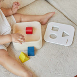 Child playing with the PlanToys shape and sort it out bin wooden shape sorter toy. Square box with lid with three shaped holes in the lid. Yellow triangle, red cylinder and blue cube blocks.
