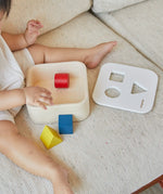 Child playing with the PlanToys shape and sort it out bin wooden shape sorter toy. Square box with lid with three shaped holes in the lid. Yellow triangle, red cylinder and blue cube blocks.