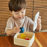 Child playing with the PlanToys shape and sort it out bin wooden shape sorter toy, lifting the lid and placing the coloured wooden blocks inside.