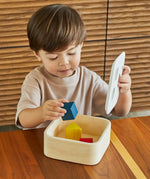 Child playing with the PlanToys shape and sort it out bin wooden shape sorter toy, lifting the lid and placing the coloured wooden blocks inside.