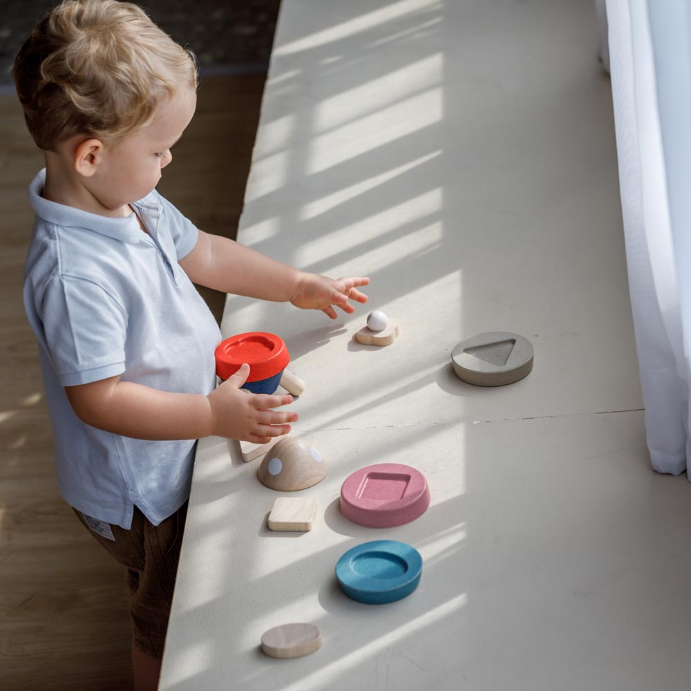 A child playing with the PlanToys Stacking Rocket Baby Toy, the pieces of the rocket have been taken off the base and have been placed all over a counter. 