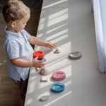 A child playing with the PlanToys Stacking Rocket Baby Toy, the pieces of the rocket have been taken off the base and have been placed all over a counter. 