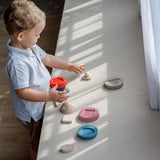 A child playing with the PlanToys Stacking Rocket Baby Toy, the pieces of the rocket have been taken off the base and have been placed all over a counter. 
