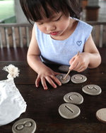 Cartoon hand reaching into a white drawstring bag filled with the Plan Toys eco-friendly wooden stone discs on a white background