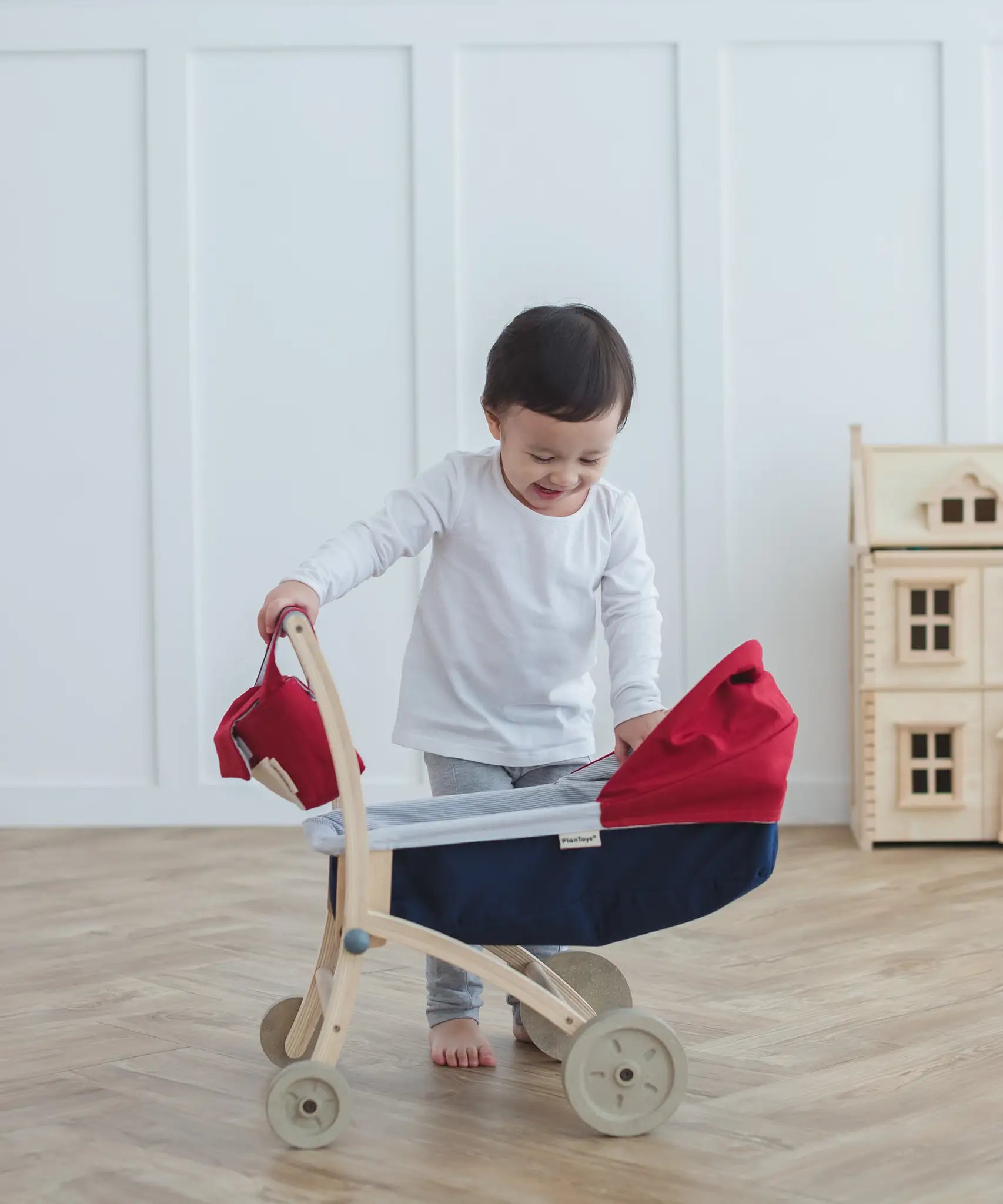Child standing next to a PlanToys stroller with a doll house in the background