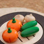 The PlanToys Vegetable Produce Set with Cucumbers, Onions & Pumpkin play food placed on a wooden toy grill. 