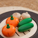 The PlanToys Vegetable Produce Set with Cucumbers, Onions & Pumpkin play food placed on a wooden toy grill. 