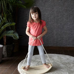 A child on the PlanToys Balance Board holding onto the rope attached to both sides of the board. The board has been placed on a circular shaped beige coloured rug. 