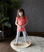 A child on the PlanToys Balance Board holding onto the rope attached to both sides of the board. The board has been placed on a circular shaped beige coloured rug. 