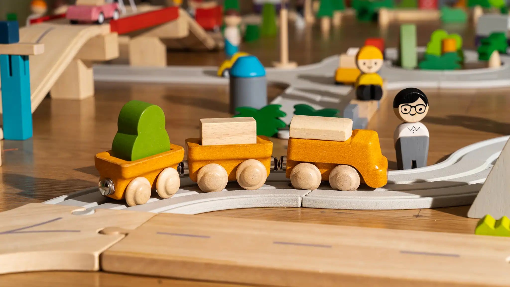 PlanToys yellow wooden cargo train placed on a rubber railway track connected to a wooden road set. 