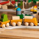 PlanToys yellow wooden cargo train placed on a rubber railway track connected to a wooden road set. 
