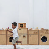A row of PlanToys wooden play kitchen units against a wall with a child running past