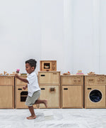 A row of PlanToys wooden play kitchen units against a wall with a child running past