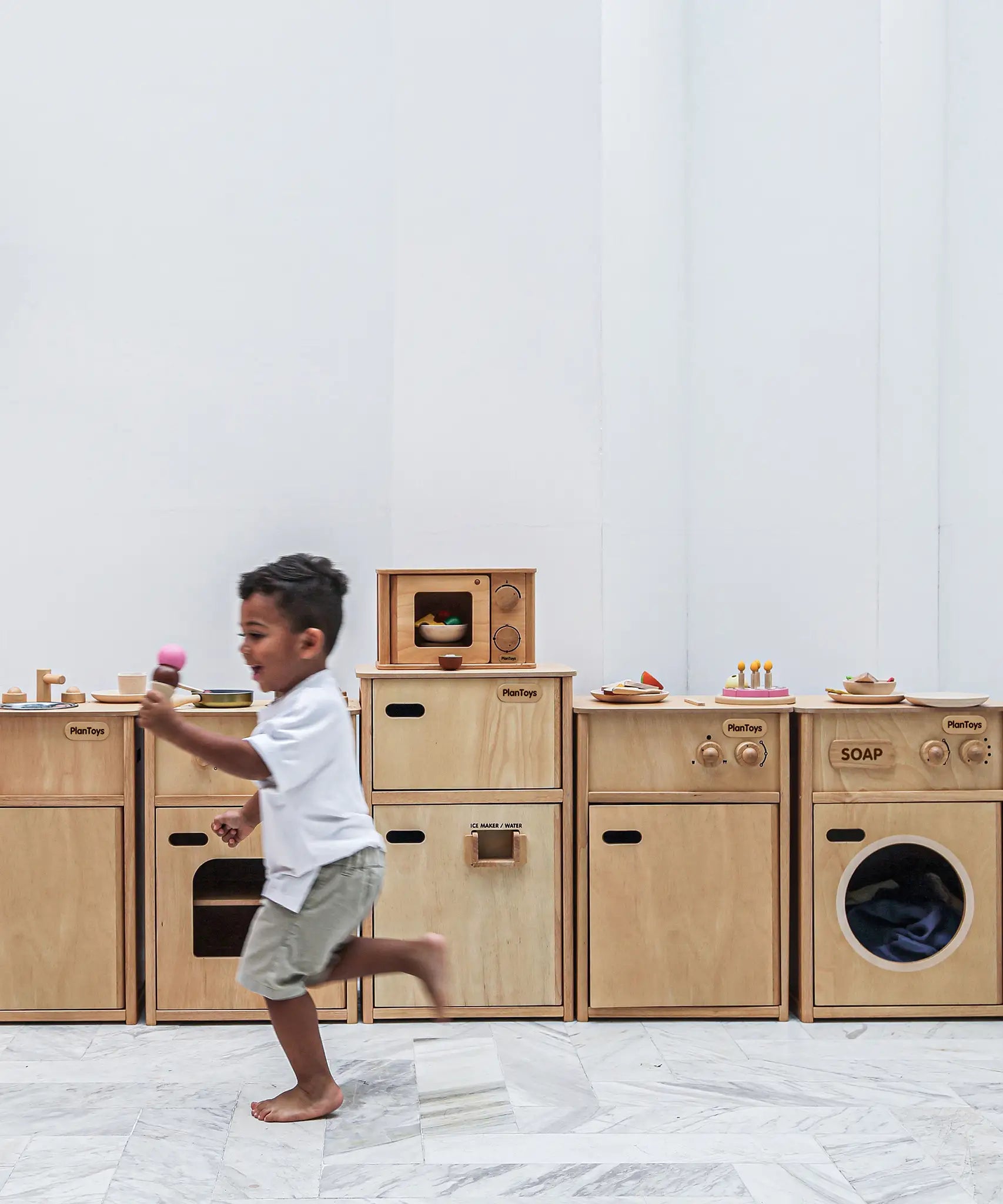 A row of PlanToys wooden play kitchen units against a wall with a child running past