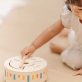 Close up of a child hand placing the wooden drumstick on top of the PlanToys wooden sensory drum musical toy.